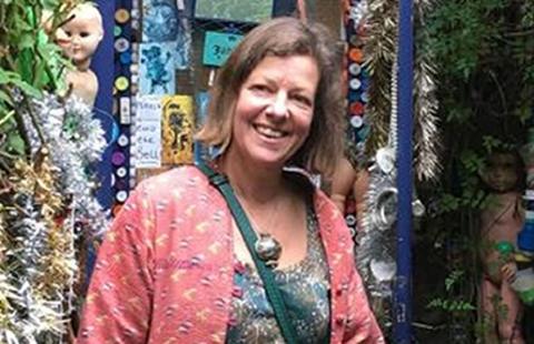 A woman with a short brown bob, a patterned pink jacket and turquoise top smiles at the camera in front of a shop front. 