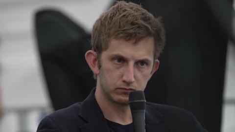 Owain Williams wearing a black jacket and holding a microphone to his face