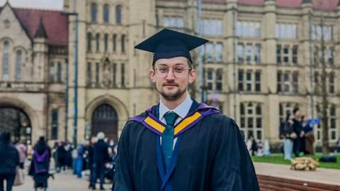 Chris Bartley, a man with brown hair and stubble, he wears glasses and a graduation cap and gown in front of a large university building.