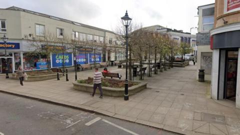 Commercial Square in Camborne. People are walking and sitting walking in the paved square, which features stone planters and benches. Black lamp posts and trees also line it. 