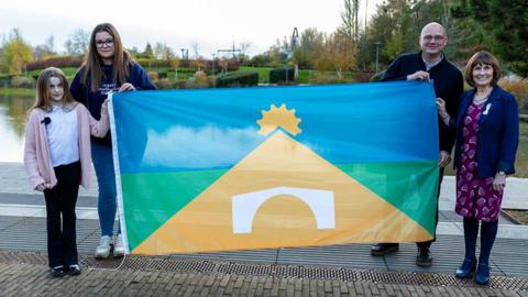 A girl wearing a white T-shit and pink cardigan and a woman wearing glasses and a navy blue hoodie are standing on one side of a flag, while a man wearing a navy blue top and a woman wearing a purple dress and navy blue jacket are standing on the other side. The flag features a design comprising yellow, blue and green elements.