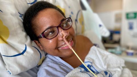 A woman lies in a hospital bed with an oxygen tube in her nose, she is wearing a hospital gown and glasses. She is looking off camera and smiling. 