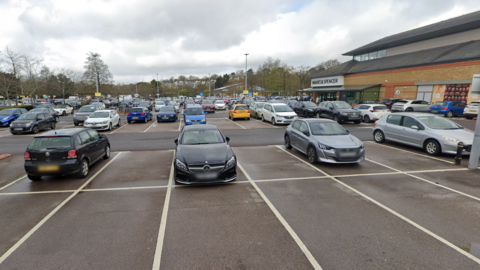 An out of town shopping centre car park which is around half full. It is bordered by a brown shop building with the sign reading Marks and Spencer.