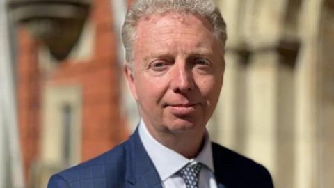 Alex Allinson, who has short wavy light coloured hair and is wearing a blue jacket and tie with a white shirt.