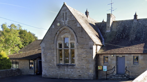 The exterior of Startforth Community Hall. It is made out of stone and has a large arched window on the side of the main hall. A blue door on the right of the building has black railings on either side of the entrance.