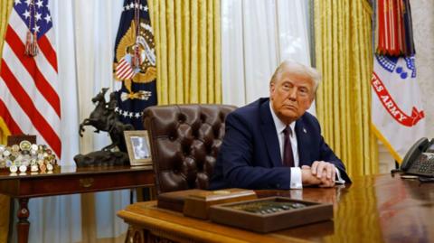 President Donald Trump sits at his desk in the Oval Office as he speaks to reporters after signing a series of executive orders
