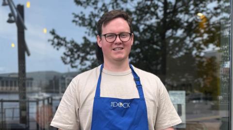 Stephen McIlwaine with short brown hair and black circular glasses on. 
He is wearing a blue apron with 'DOCK' written on it and a cream t-shirt.
He is standing in front of a glass window, smiling at the camera. 