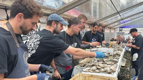 A line of men in black T-shirts shuck oysters