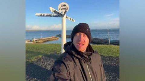 Jack Ward is in a black waterproof jacket and black beanie. He is taking a selfie in front of a white sign for John O'Groats. There is grass behind him, and beyond that is the sea.