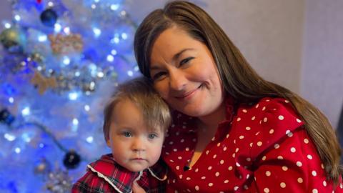  A woman wearing a red polka-dot blouse is sitting indoors in front of a decorated Christmas tree, holding a young child dressed in a red tartan outfit.