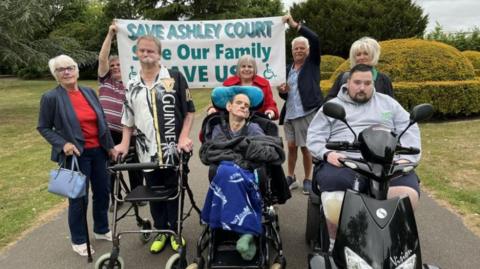 A group of eight people with two men at the back holding up a sign that reads 'Save Ashley Court'. One person at the front has a zimmer frame, another is in a wheelchair and another is on a mobility scooter.