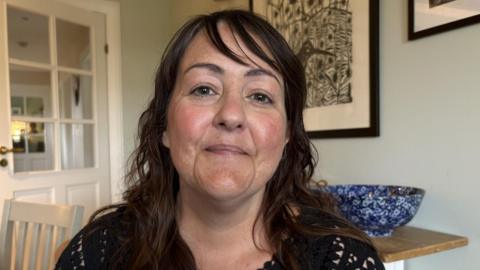 Kelly Davison has shoulder-length, dark hair. She is sitting at a kitchen table and smiling at the camera. She is wearing a black top. There are pictures behind her and a door off to the left. 