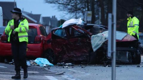 Crash scene shows two badly damaged cars and debris in the road. Two police officers wearing yellow hi-vis jackets are at the scene.
