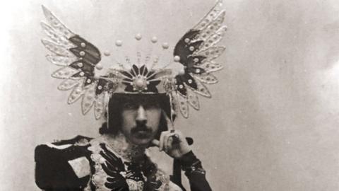 The 5th Marquess of Anglesey in a sepia photograph wearing a floor length overcoat consisting of differently coloured pieces of heavy material. The top of the coat is military in style with large shoulder pieces. He wears a headdress of splayed wings with a tiara-effect frontspiece.