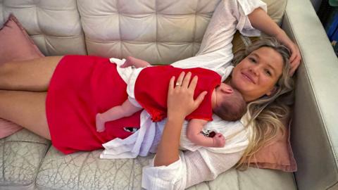 A woman with long blonde hair, wearing a white top and red skirt lying on a cream couch, with a baby sleeping on her chest.