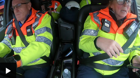 Internal car view of a male traffic police officer driving on the right and female officer on the left looking out of the window. They are wearing yellow hi-vis uniform.