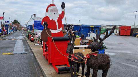 Father Christmas wearing a red, white and black outfit sits on the back of a sleigh made out of household waste. It looks like it is being pulled by two reindeer which are also made out of rubbish. The sculpture sits on a concrete platform next to a road at Grimsby tip. Waste bins and other containers are visible in the background.