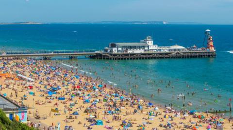 Bournemouth beach, a sandy beach which is packed with hundreds of people, tents and umbrellas. Bournemouth pier is in the background.