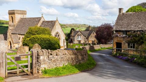 The Cotswolds village of Snowshill, Gloucestershire, on a sunny day. A country lane bordered by a traditional Cotswold Stone wall meanders past a church, a house, and a pub. Rolling hills with woodland on them can be seen in the distance.