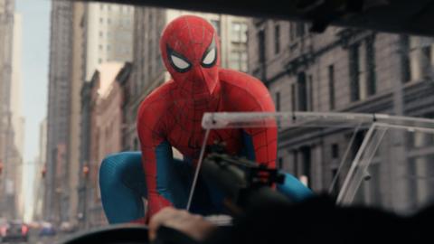 Spiderman on the bonnet of a car, with a number of tall stone buildings behind him. 