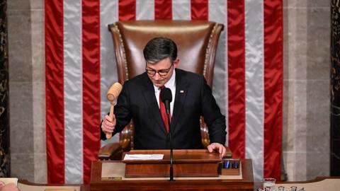 Mike Johnson wearing a black suit, red tie and slams a gavel in front of an American flag