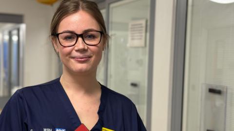 Senior Sister Charlotte Moore stands in the emergency department. She has brown hair that is tied back and is wearing glasses. She is also wearing a dark blue uniform.