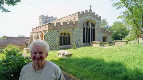 Ewelme historian Carol Sawbridge smiling for a picture in front of Ewelme Church. It is a sunny day.