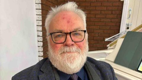 Alan Gregory, piano teacher, sits at a white piano in a dark three piece grey and navy suit. He has grey hair, a grey beard, blue eyes and wears glasses.