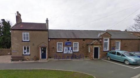 A brown brick school building, with cars parked in bays located in front. To the left is a small section of grass. 