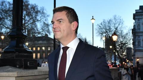 Olly Robbins walking down Whitehall in central London wearing a navy suit and a red tie