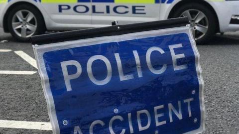 A blue rectangular sign with white lettering saying "police accident" resting on a road. Behind it is the lower part of the side of a police car in yellow, blue and white livery.