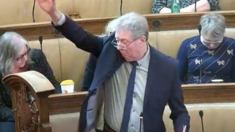 A screengrab taken from a live stream of a Hull City Council meeting. Alan Gardiner, wearing a dark blue suit, white shirt and blue tie is standing with his arm in the air. He has grey hair and glasses.