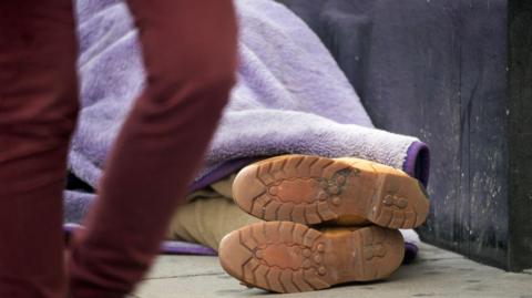A person lies on the pavement wrapped in a purple blanket, with only their boots visible, as someone walks past in the foreground.