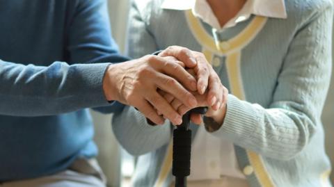 generic image of an anonymous elderly couple sitting together and clasping hands over the lady's walking stick