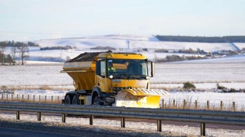 A yellow snow plough gritter on a road with snowy fields and hills in the background.