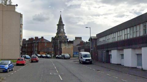 A street in Glasgow with a commercial building on one side and flats on the other. Cars are parked at the side of the road and a spire is seen at the far end of the street.