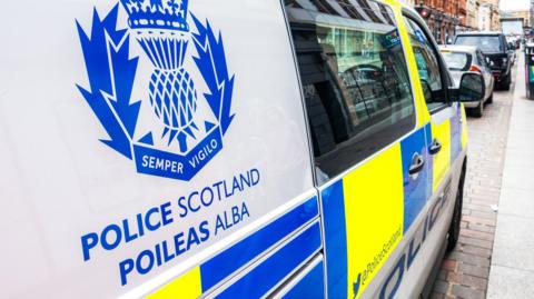 A close up of a blue Police Scotland logo printed onto a police van. The logo is a thistle with the words Semper Vigilo written underneath. Below that are the words Police Scotland and Poileas Alba. The van is parked at the side of a street.