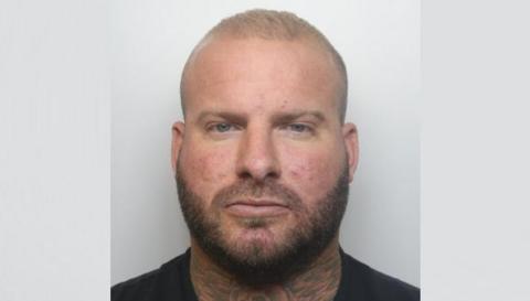 A mugshot of Alex Male standing against a grey wall. He is wearing a black t-shirt and short blonde hair.