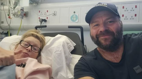 Stacey, on the left in a hospital bed, giving a thumbs up sign while Stu, wearing a basaeball cap, takes a selfie of them both 