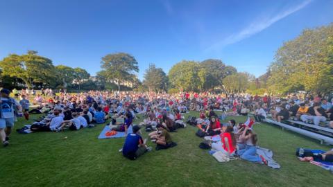 Hundreds of people sit on the grass of a park on a sunny day.