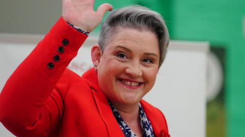 Labour MP for West Lancashire Ashley Dalton with short grey hair wearing red lipstick, an emerald studded earring with a second hoop earring smiling and waving. She is wearing a blue and white patterned blouse and bright red jacket. 