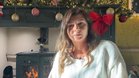 Agnieszka McIntyre has shoulder-length brown hair and is wearing a white knitted jumper. She sits in front of a black metal wood-burning stove below a black fireplace decorated with Christmas baubles and red Christmas bows.
