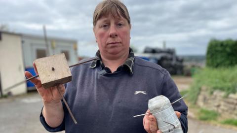 A middle-aged woman with hair tied back and a navy jumper holds up a block of wood and a small bottle. Both have sharp nails poking out.