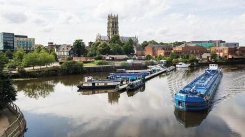The photo shows a wide stretch of river with a blue barge in the forefront and a number of smaller barges in the background. Doncaster Minister overlooks the water