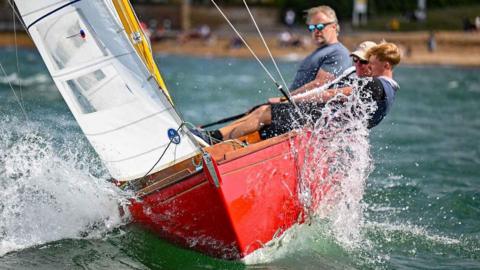 Three people sailing a Magic Dragon XOD class sailing boat. The boat is red with a white sail and the people are leaning back as the boat tilts.
