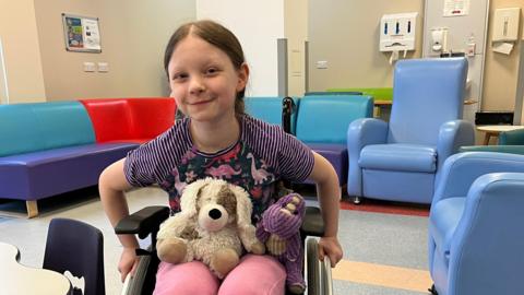 Ivy is sitting in a wheelchair in what looks like a hospital waiting area and smiling at the camera. Two teddy bears sit on her lap. Behind her is a multi-coloured L-shaped vinyl sofa and a couple of blue very high-back armchairs.