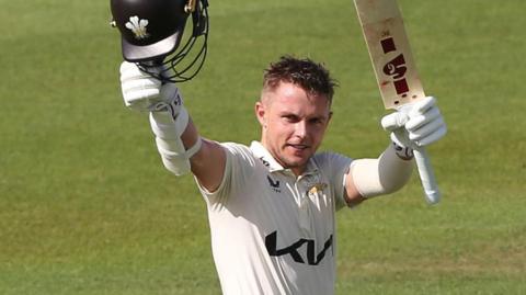 Sam Curran raises his bat and helmet after reaching 100 for Surrey against Durham