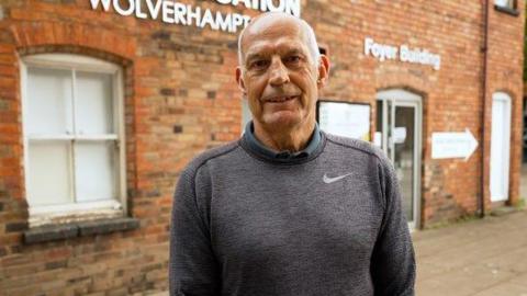 A man with short grey hair on the sides of his head is looking at the camera and smiling. He is wearing a grey Nike jumper and is standing in front of a red brick building.
