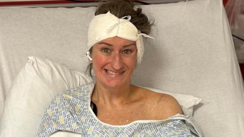 Susannah is propped up in a hospital bed with a bandage around her head. She is wearing a hospital gown and is attempting to smile but one side of her mouth is drooping down.