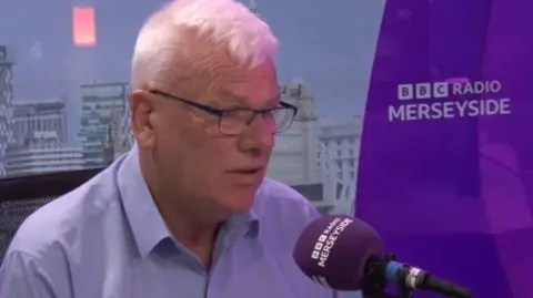 Graham Morgan, wearing glasses and a blue shirt, sits in a purple BBC Radio Merseyside studio